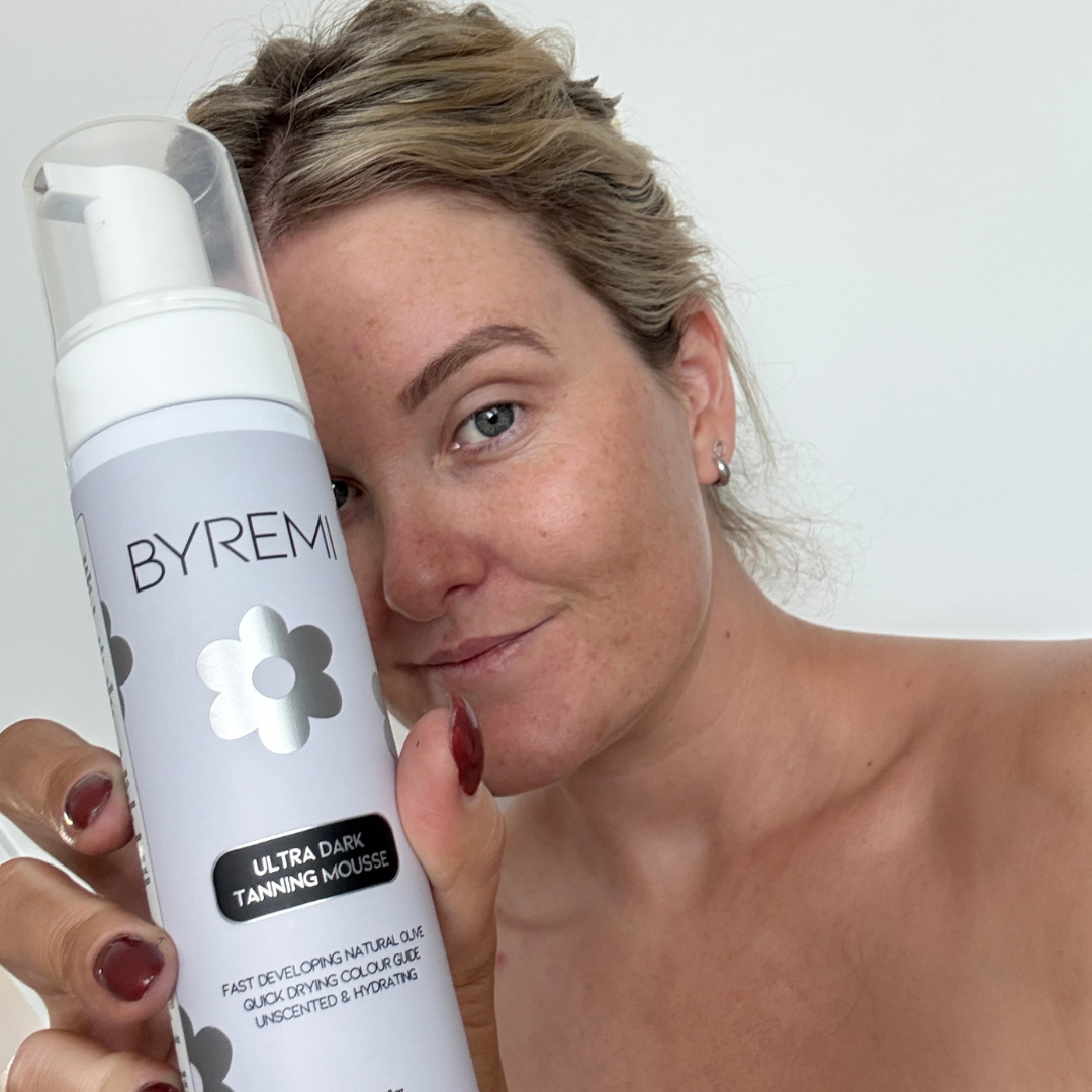 Woman holding a Byrem ultra dark tanning mousse bottle against a white background