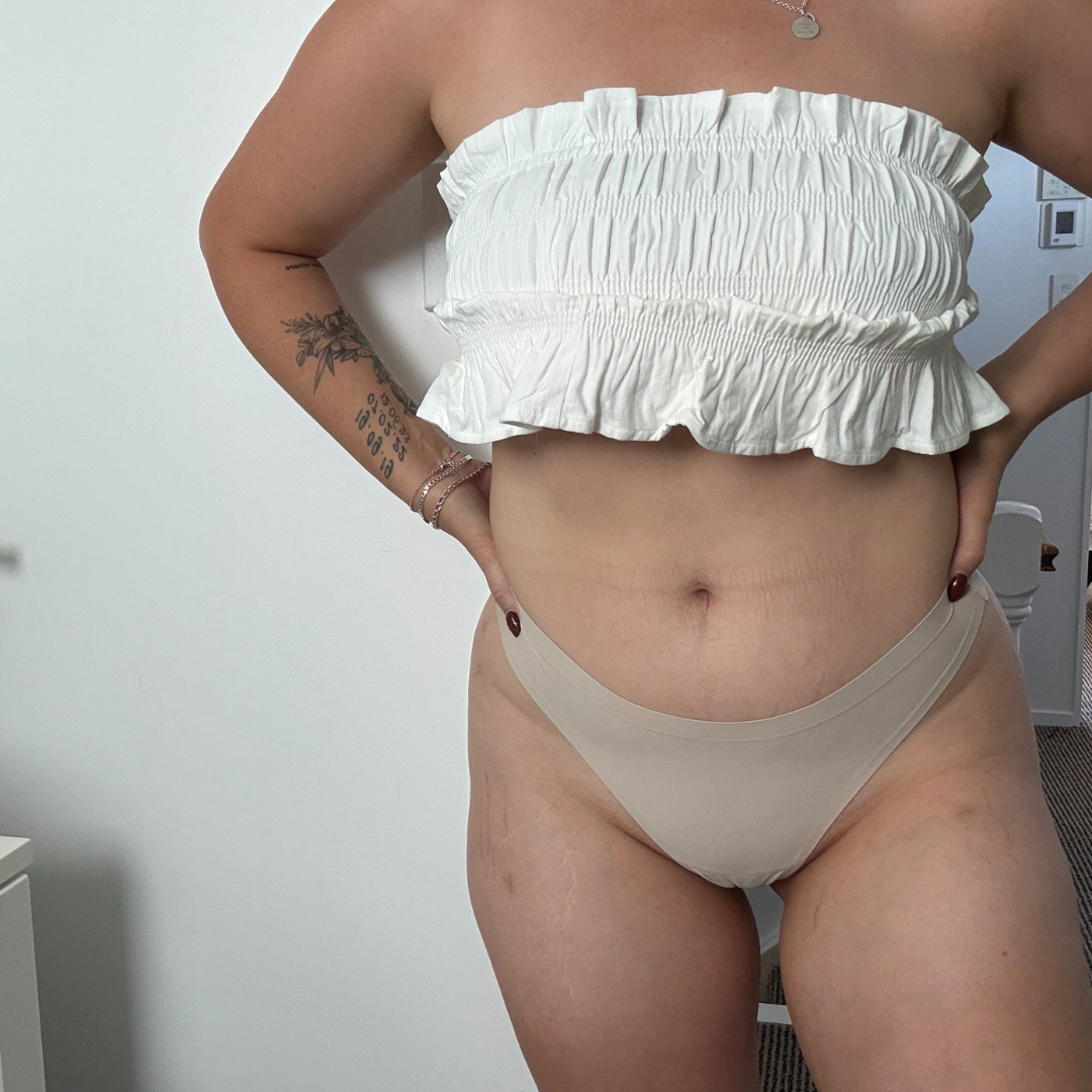 Person wearing a white ruffled top and beige bikini bottom against a neutral background self tanned