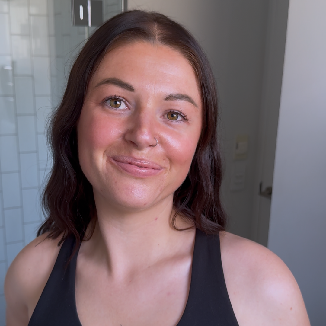 Woman with brown hair and a black tank top standing in a bathroom showing self tan